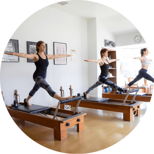 Three women in athletic wear perform Pilates on reformer machines in a bright studio, arms extended forward in a synchronized pose.